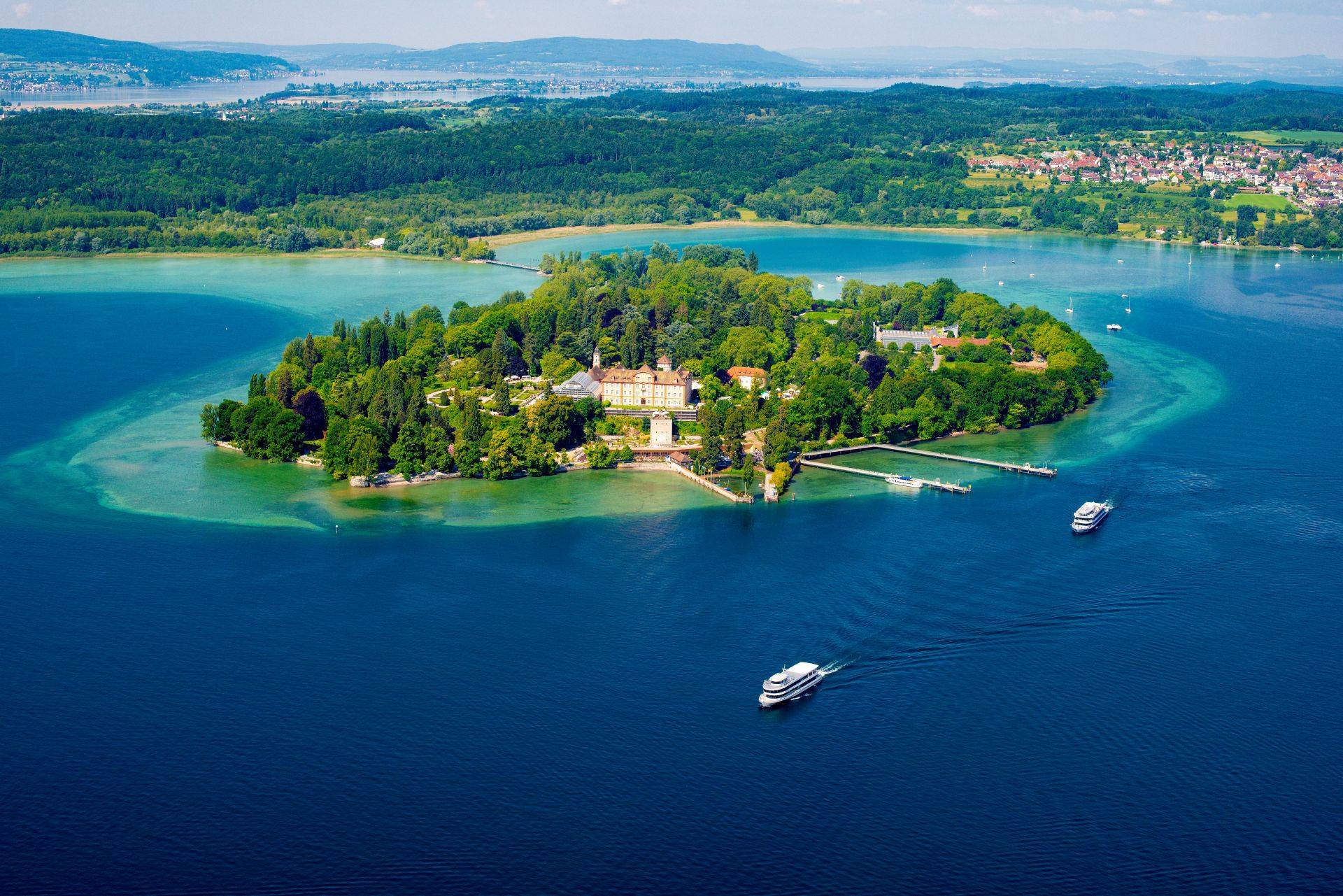 Luftaufnahme der Insel Mainau im Bodensee mit Schloss und Ausflugsbooten, umgeben von klarem Wasser und grüner Landschaft